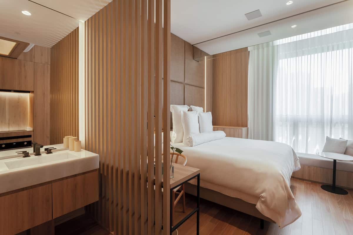 A modern hotel room with a wooden panel wall, a large bed with white linens, a round table, and a large window with sheer curtains.