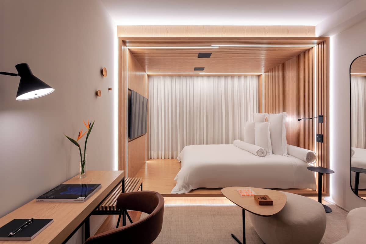 A modern hotel room with a bed, a desk, a chair, a small table, and a TV.