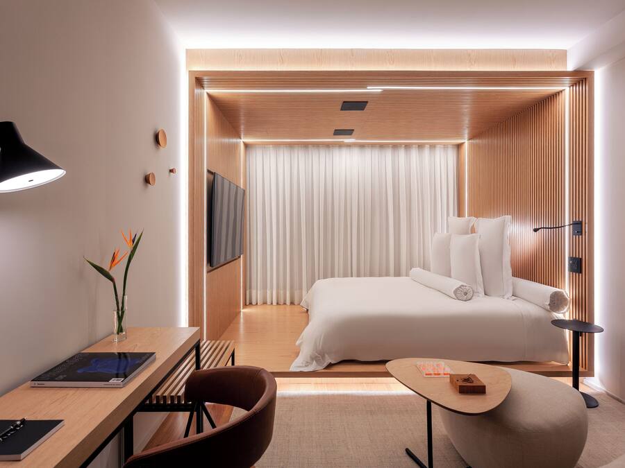 Quarto de hotel moderno com cama, escrivaninha, cadeira, mesinha e televisão.