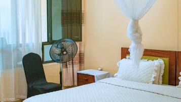 A hotel room with a bed, a standing fan, a chair, a nightstand, and a window with curtains.