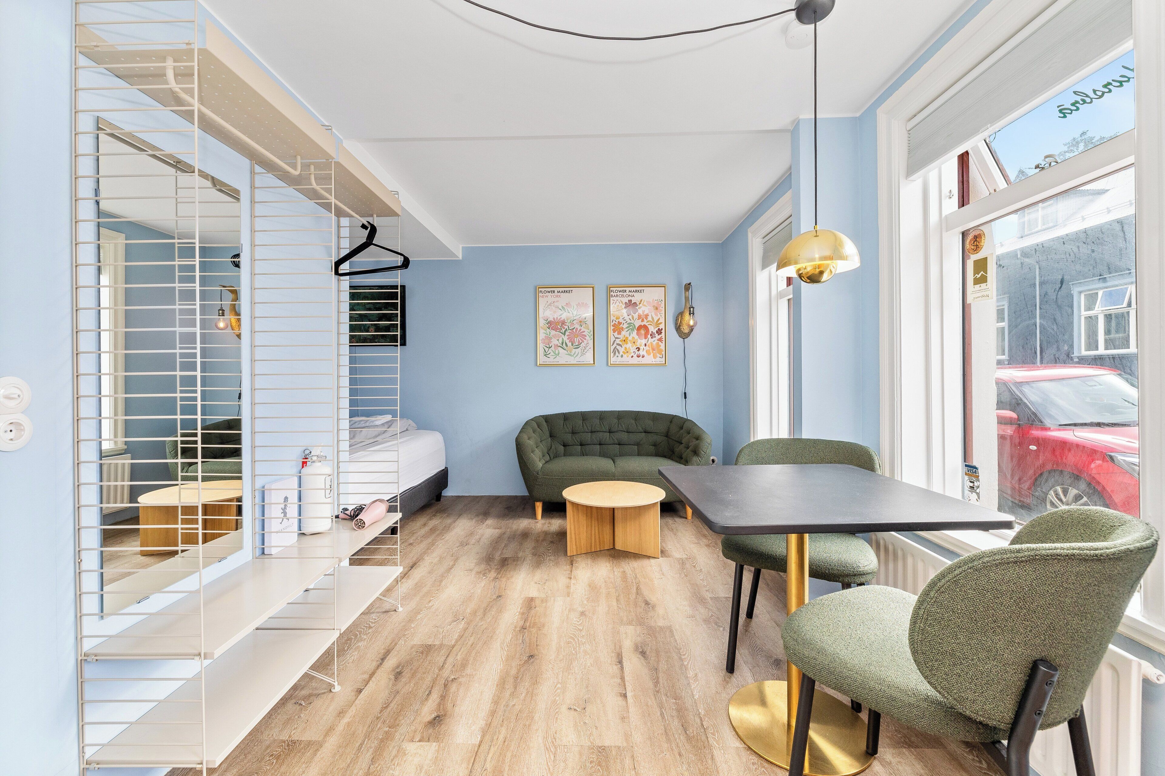 A modern room with a bed, a green sofa, a round wooden table, and chairs. There are two framed pictures on the wall, a hanging light fixture, and a window with a view of the outside.