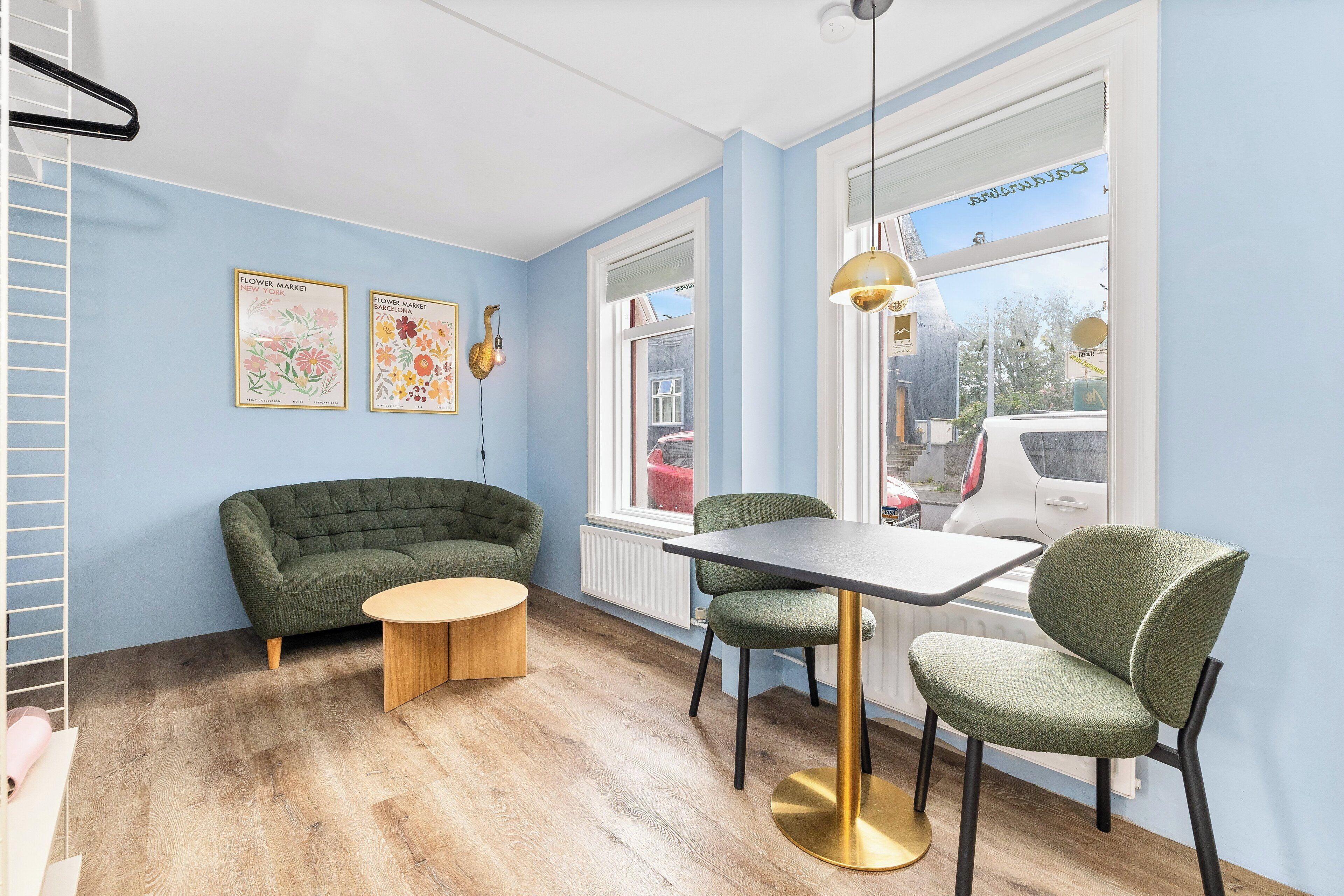 Ein Zimmer mit einem grünen Sofa, einem runden Holztisch und zwei grünen Stühlen. An der Wand hängen zwei gerahmte Blumenbilder und durch das Fenster ist ein Blick auf parkende Autos zu sehen.