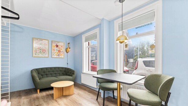 A room with a green sofa, a round wooden table, and two green chairs. There are two framed floral posters on the wall and a window with a view of parked cars outside.
