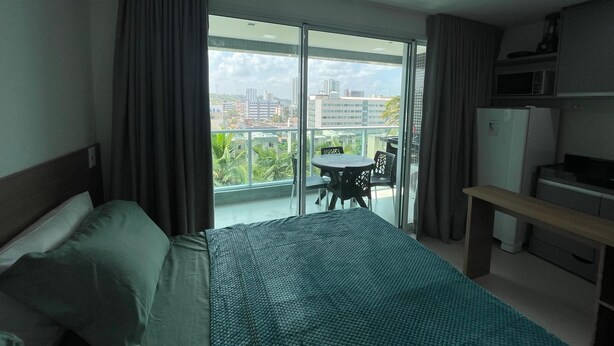 A hotel room with a bed, a small table with chairs, a refrigerator, and a view of a cityscape through a large sliding glass door. - RN Studio Premium 503 - Vista Mar (Maceió)