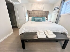 A neatly made bed with a tufted headboard, a bench with folded towels, and a door leading to another room. - Spacious 6-Bedroom, 2.5-Bath Home Near Downtown w/ 4 Parking Spots and A/C (Vancouver)