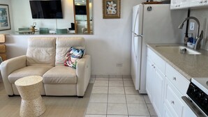 Een moderne keuken met witte kastjes, een inbouwoven en een spoelbak. Er staat een beige leren bank met een tropisch kussen en een geweven kruk. Een plafondventilator en een lijst met een afbeelding hangen aan de muur.