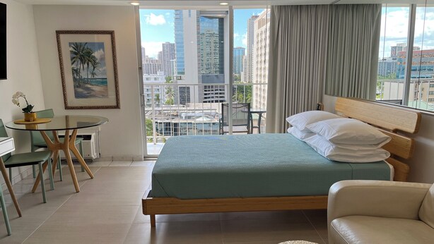 Een moderne hotelkamer met een groot bed, een bank, een eettafel met stoelen en uitzicht op de skyline door grote ramen.