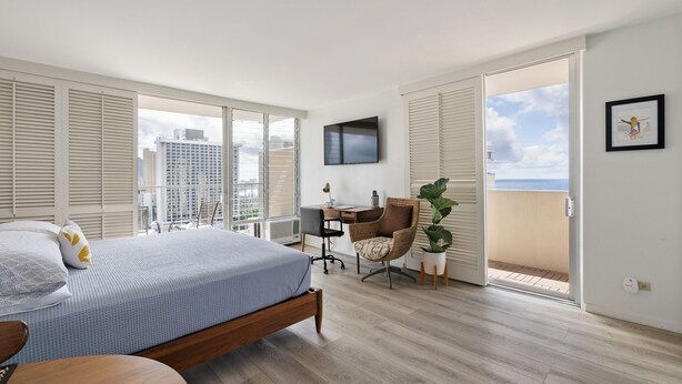 A modern bedroom with a large bed, a desk, and a chair. There is a TV mounted on the wall and a balcony with a view of the city. - Dazzling Waikiki Studio+KIng Bed (Honolulu)