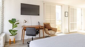 A hotel room with a bed, a desk with a chair, a television, and a large window with blinds. - Dazzling Waikiki Studio+KIng Bed (Honolulu)