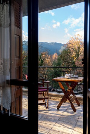 Un balcon avec une table et des chaises, une vue sur les arbres et les collines, et une fenêtre avec des rideaux.