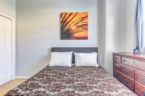 Bed with brown and blue patterned quilt, gray upholstered headboard, wooden dresser, and sunset palm print above.