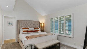 Un dormitorio con cabecera capitonada, una cama grande con almohadas a rayas, un banco, una mesita de noche y una ventana con persianas.