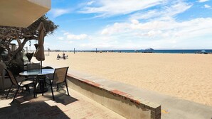 Blick auf einen Strand mit einem Tisch und Stühlen unter einem Sonnenschirm, eine Ziegelmauer und ein klarer Himmel.