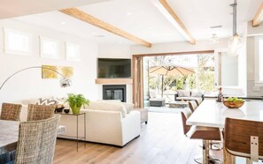 A modern living room with a fireplace, a dining area, and a large window overlooking a garden.