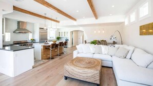 A modern kitchen with a central island, a dining area with bar stools, a white sofa, and a woven ottoman.