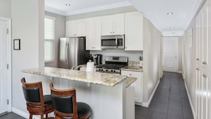 A kitchen with white cabinets, a granite countertop, stainless steel appliances, and a dining area with two chairs. - Newport Beach Retreat Steps to Sand, Boardwalk and Pier (Newport Beach)