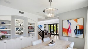 Un espace repas moderne avec une table en bois clair, des chaises blanches et un lustre. On y trouve des meubles blancs ornés d’objets décoratifs et un grand tableau abstrait accroché au mur.
