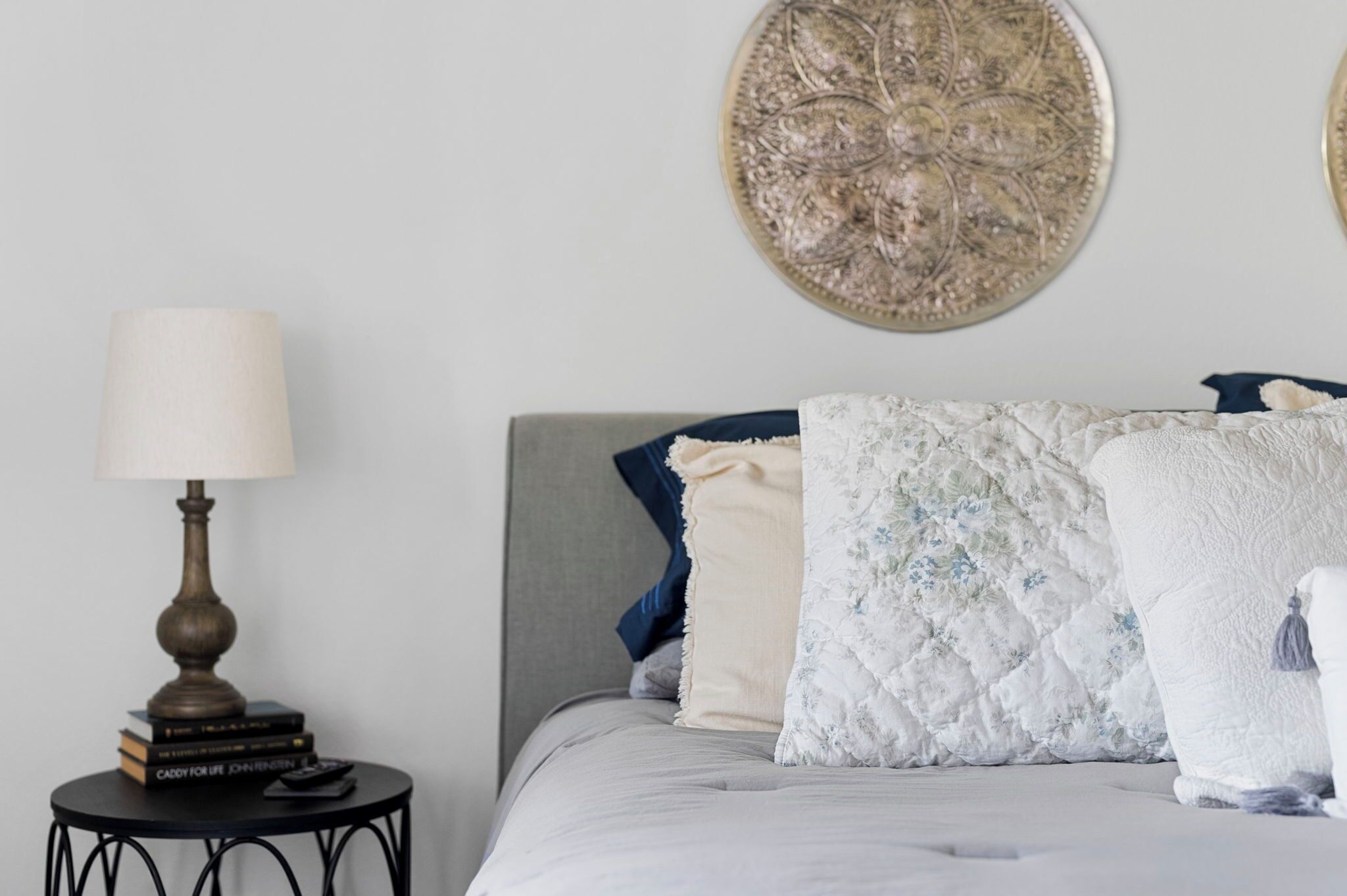 A neatly made bed with a grey headboard, a bedside table with a lamp and books, and decorative pillows.