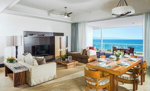 Relax in a spacious living area with a large flat-screen TV, comfortable sofa, and dining table, plus a balcony with ocean views.