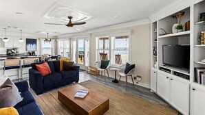 Amplia sala de estar con sofá, mesa de centro y sillas, una cocina con barra y un televisor sobre un mueble blanco.
