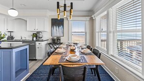 Una cocina moderna con armarios blancos, un comedor con mesa de madera dispuesta para seis comensales y ventanas amplias con persianas.