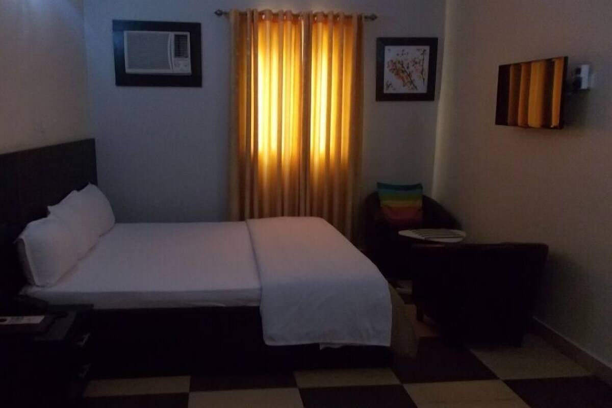 Quarto de hotel com uma cama, mesa de cabeceira, aparelho de ar condicionado, janela com cortinas e um quadro pendurado na parede.