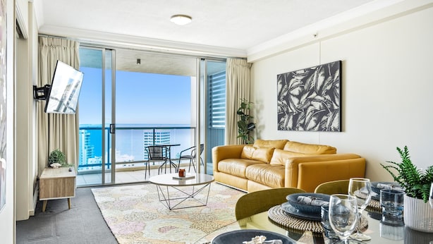 A modern living room with a sofa, a dining table set with plates and glasses, and a view of the ocean through large glass doors. - 2 Bedroom 2 Bathroom Ocean View Unit at Chevron (CHEV 3204 - 3N - NB) (Surfers Paradise)