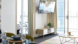 A modern hotel room with a dining area, a flat-screen TV mounted on the wall, a small wooden side table, and a large window offering a city view. - 2 Bedroom 2 Bathroom Ocean View Unit at Chevron (CHEV 3204 - 3N - NB) (Surfers Paradise)