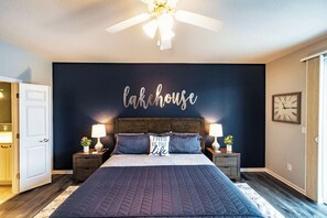 A bedroom with a dark blue wall, a bed with a quilted cover, two bedside tables with lamps, a ceiling fan, and a clock on the wall.