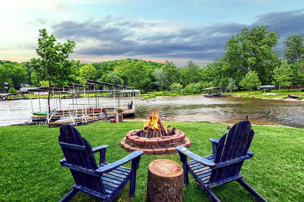A riverside deck with a fire pit, two Adirondack chairs, and a dock with boats.