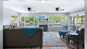 A spacious living room with a large sofa, a coffee table, and a TV. There are two ceiling fans, large windows with a view of the outdoors, and a small balcony with a railing. - FiveStar Homestead | Ozark Waterfront Living for 8 w/ Private Dock! (Camdenton)