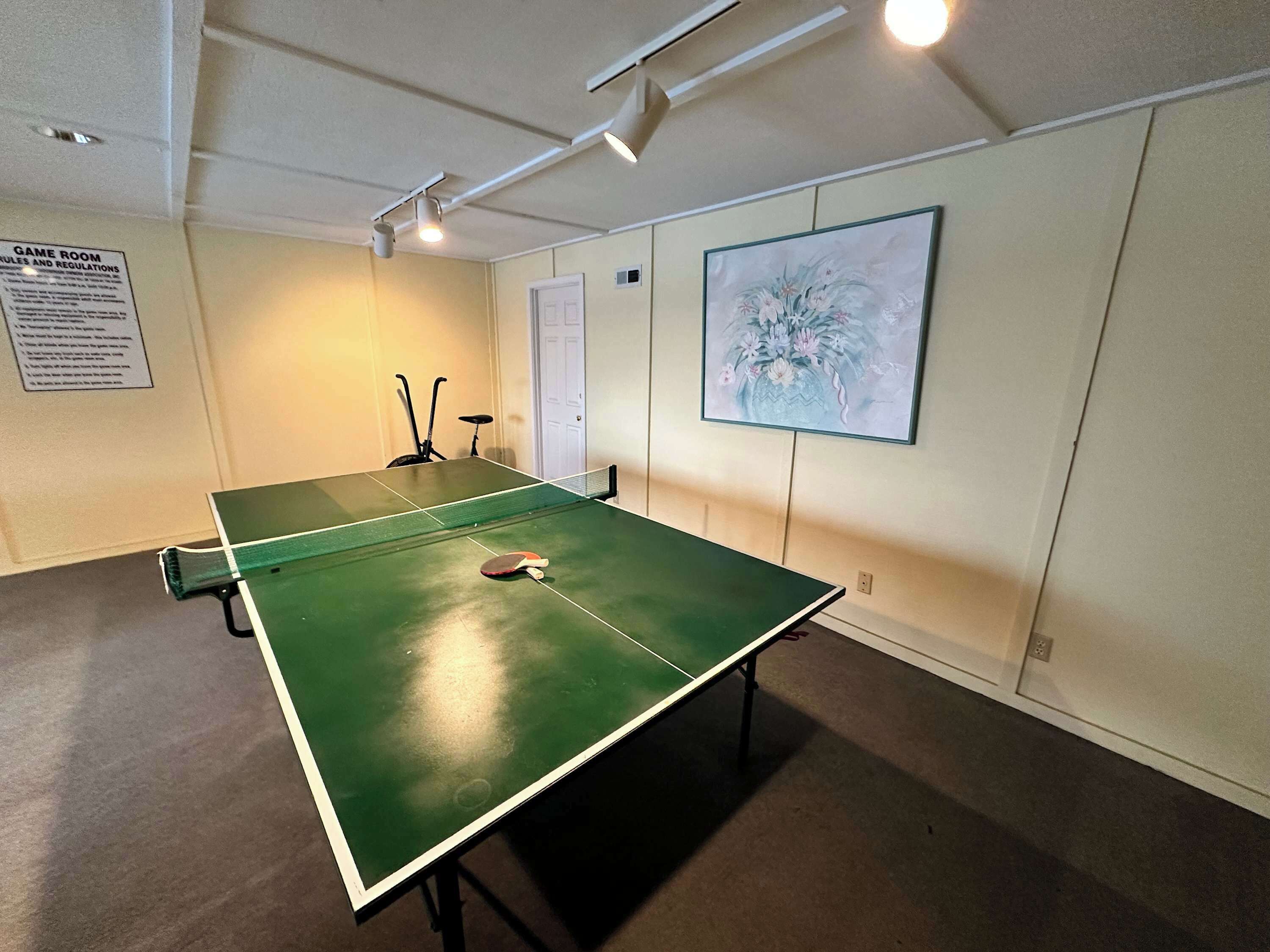 Una mesa de ping pong en una habitación con una puerta, una obra de arte enmarcada en la pared y un cartel con normas y reglamentos.