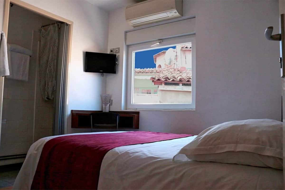 A bedroom with a bed, a TV mounted on the wall, and a window with a view of buildings.