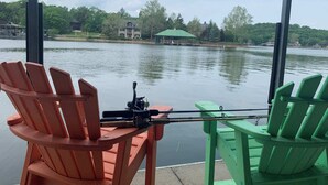 A view from a window with two Adirondack chairs, one red and one green, overlooking a calm lake with a house and a gazebo in the background. - Fairway to Heaven in Old Kinderhook | Golf (Camdenton)