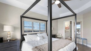 Ein Bett mit Baldachin, durch die Fenster ist Meerblick, ein Nachttisch mit Lampe und ein kleiner Stuhl in der Ecke.