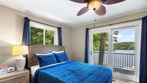 Une chambre à coucher avec un lit, une table de chevet, une lampe et une vue sur un lac, visible à travers de grandes fenêtres.