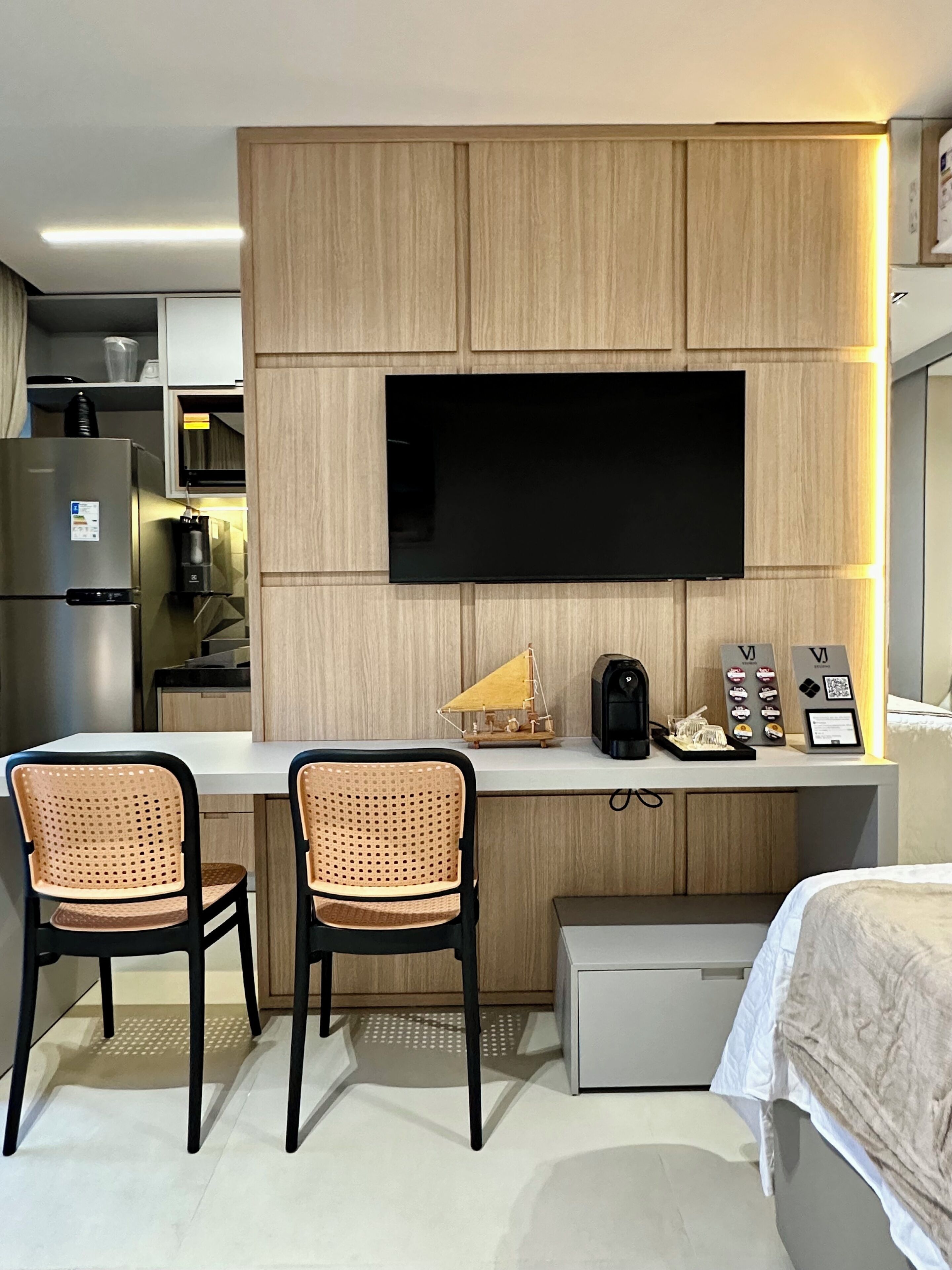 A modern hotel room with a kitchenette, a flat-screen TV mounted on a wooden panel wall, two wicker chairs, a small table with a coffee machine, and a bed with a white comforter.
