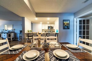 A dining area with a wooden table set for four, a kitchen in the background, and a framed picture on the wall.