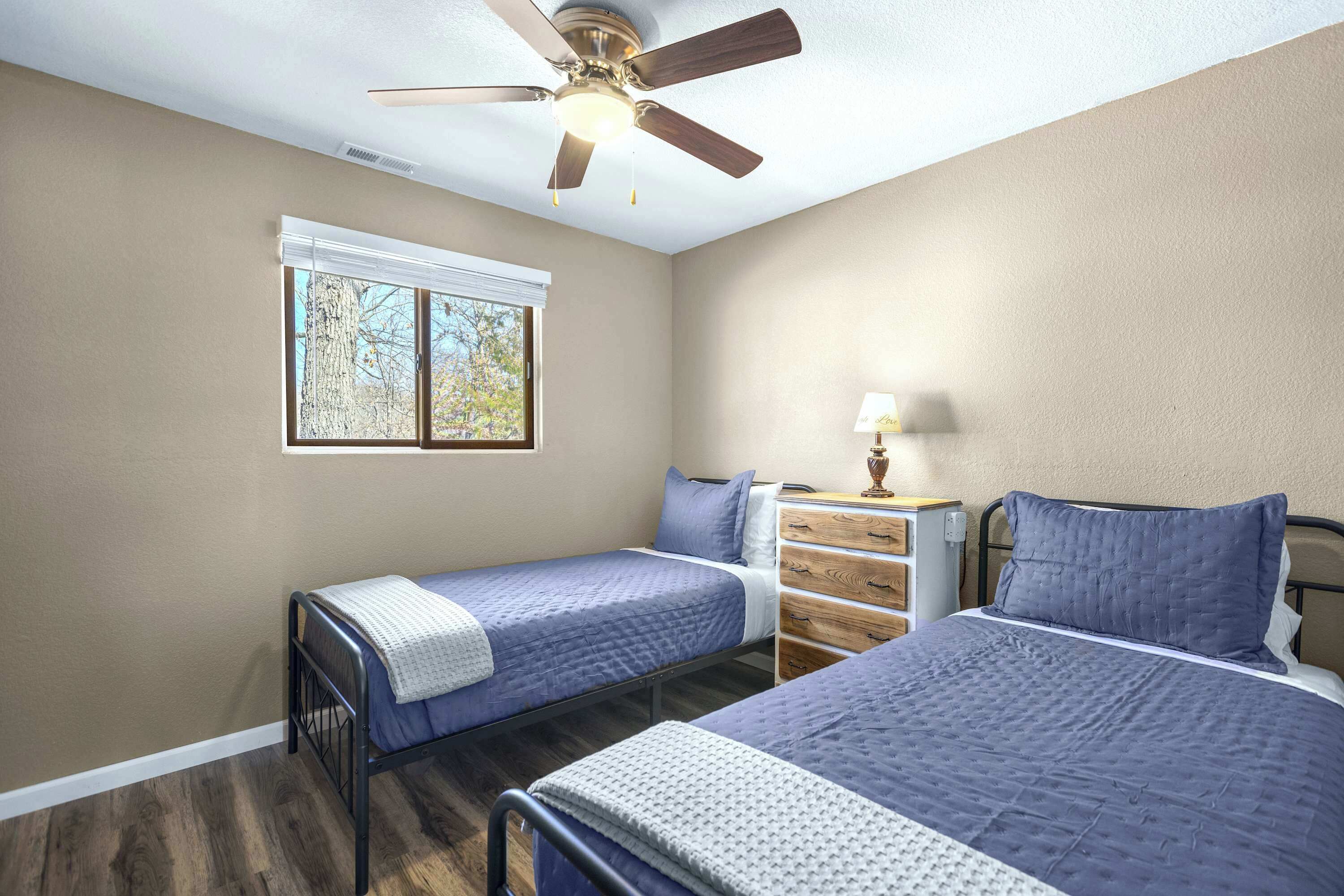 Quarto com duas camas, um ventilador de teto, uma janela com persianas, cômoda e abajur.