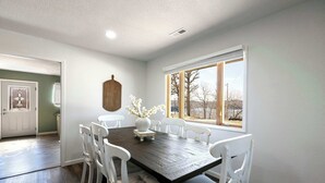Un comedor con una mesa de madera oscura y sillas blancas, un jarrón con flores y una ventana que ofrece vistas a árboles y un edificio.