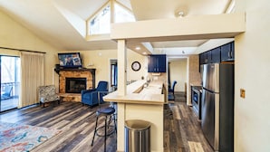Una cocina moderna con una isla central, horno empotrado y refrigerador. Hay una zona de comedor con un sofá azul y una chimenea.