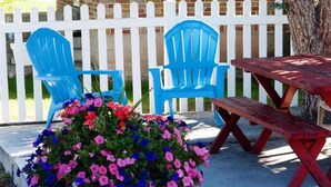A patio area with two blue chairs, a wooden table, and a flower planter with pink and blue flowers. - Blue Pine Motel (Panguitch)