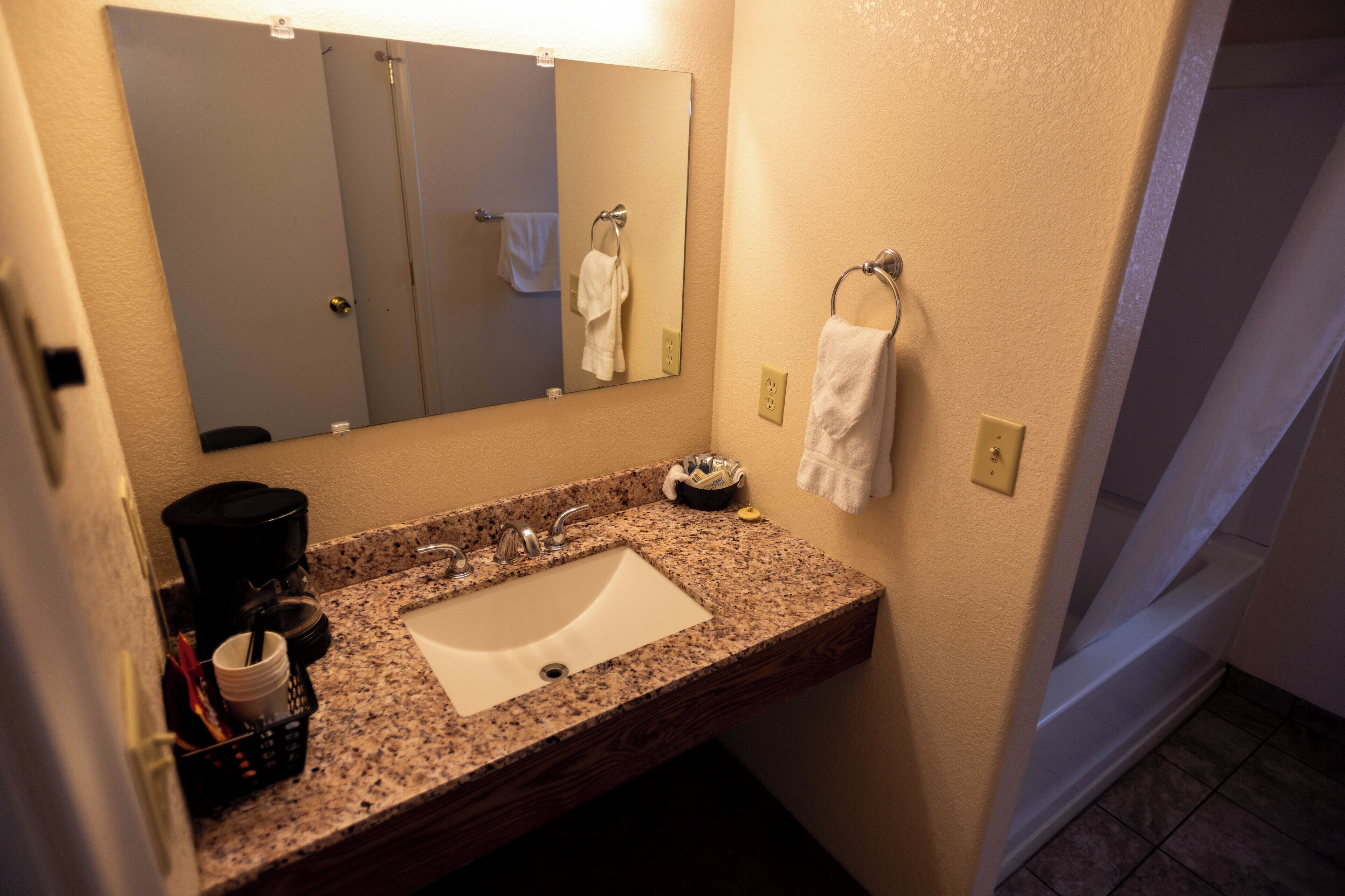 A bathroom with a granite countertop sink, a mirror, a towel ring, and a coffee maker.