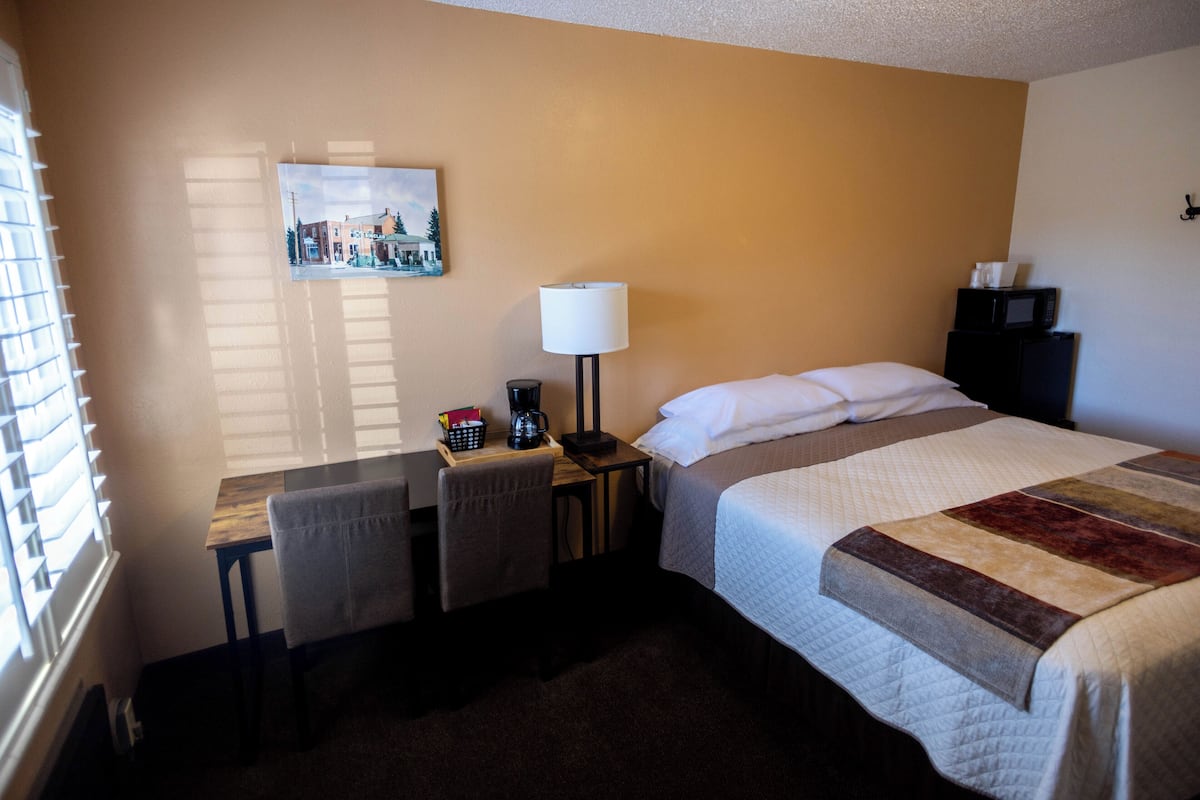 A hotel room with a bed, a desk, a chair, a lamp, a picture on the wall, and a microwave on a small table.
