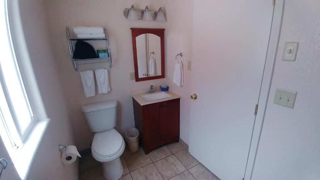 A small bathroom with a toilet, sink, and mirror. There is a shelf above the toilet holding towels and toiletries.