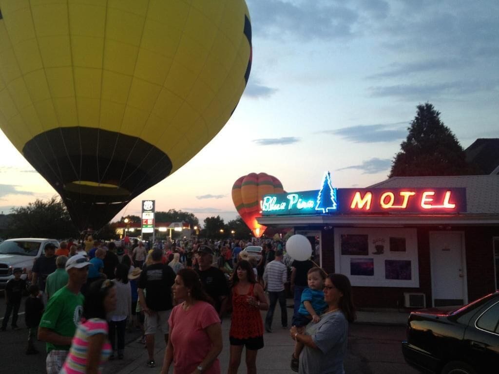 A motel with a neon sign, a hot air balloon in the background, and a crowd of people.