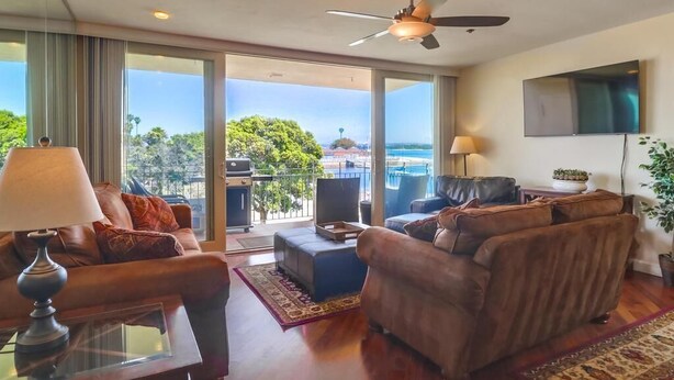 A spacious living room with a large window, a ceiling fan, and a flat-screen TV mounted on the wall.