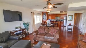 A living room with a sofa, ottoman, and a flat-screen TV. There is a ceiling fan and a kitchen in the background.