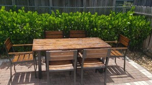 A wooden table with six chairs, set on a brick patio with a wooden fence and green foliage in the background.