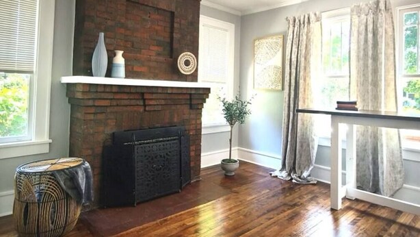 A room with a brick fireplace, wooden flooring, a ceiling fan, and a window with curtains.